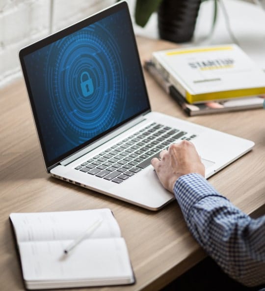 A person types on a laptop displaying a blue lock icon on its screen, with a notebook on the desk.