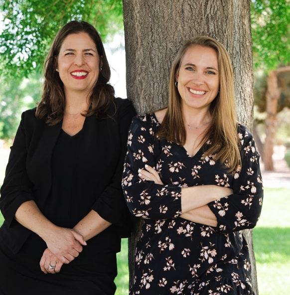 Two smiling women, one in a black suit and one in a black floral dress, lean against a tree.
