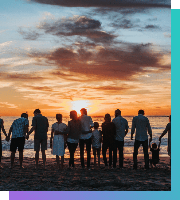 A group of people of various ages stand on a beach, silhouetted against a vibrant sunset over the ocean.